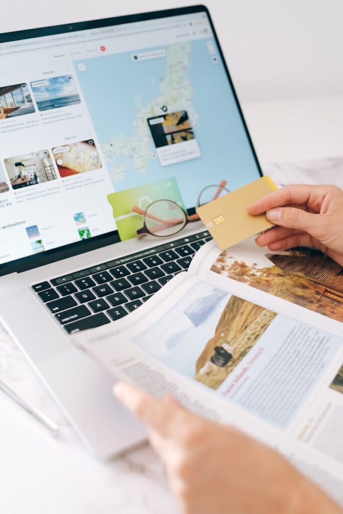 Hands holding credit card over laptop and magazine for online shopping and travel planning.